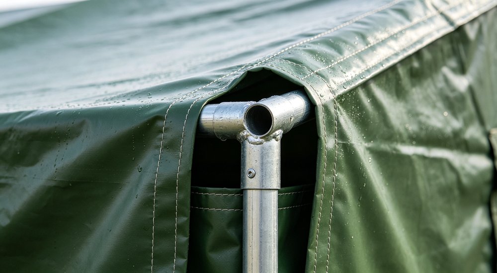 Detailed close-up shot of green relief tent metal frame structure and PVC tarpaulin fabric connection point, metal pole joint and fabric attachment with tension straps visible at tent corner showing construction quality