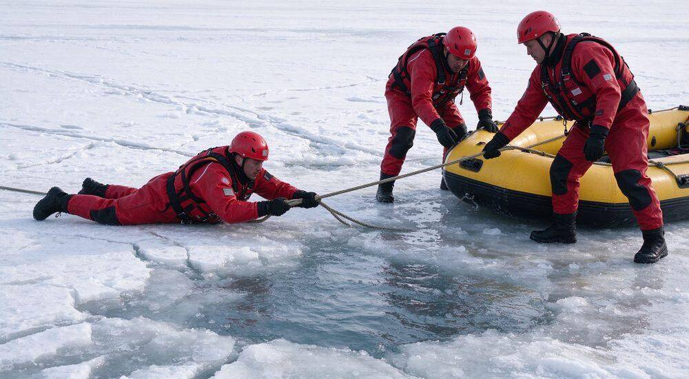 Ice rescue operation on frozen lake with three male rescuers in red drysuits and helmets pulling rescue rope near yellow inflatable rescue raft across cracked ice in cold scenario