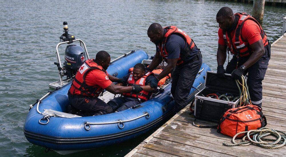 Boarding and loading operation where male rescuer stabilizes inflatable rescue boat on shore while injured victim climbs aboard showing patient transfer procedure for flood response boat