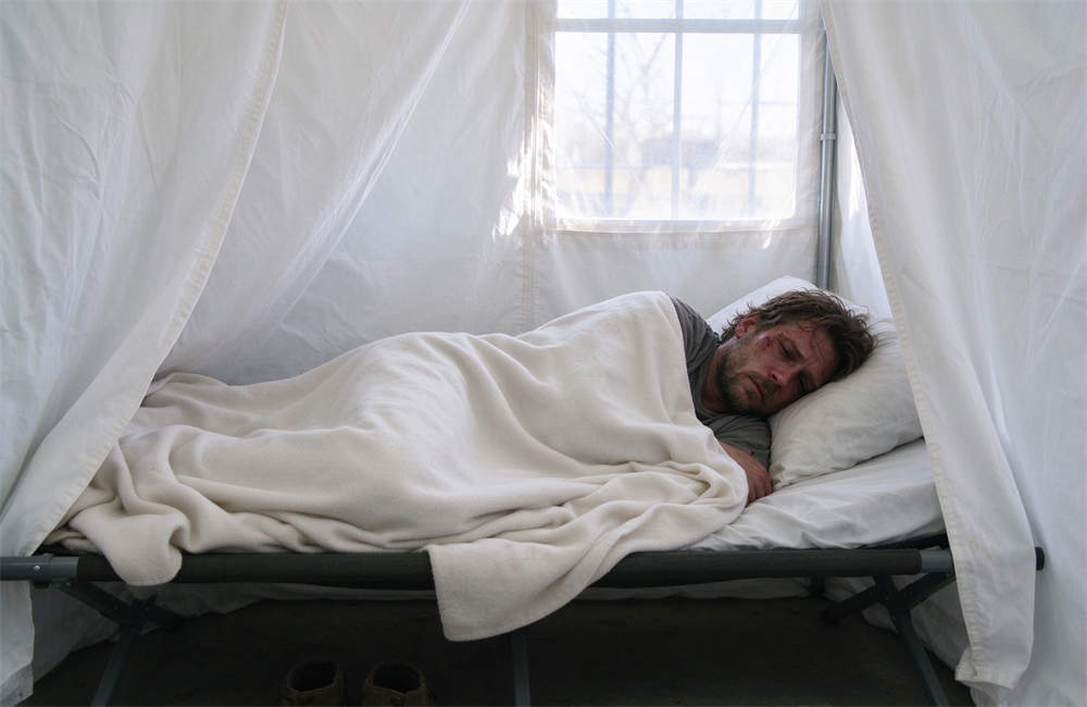 Person lying on isolation cot covered with clean white blanket, foodborne illness patient resting in relief shelter isolation area with medical monitoring